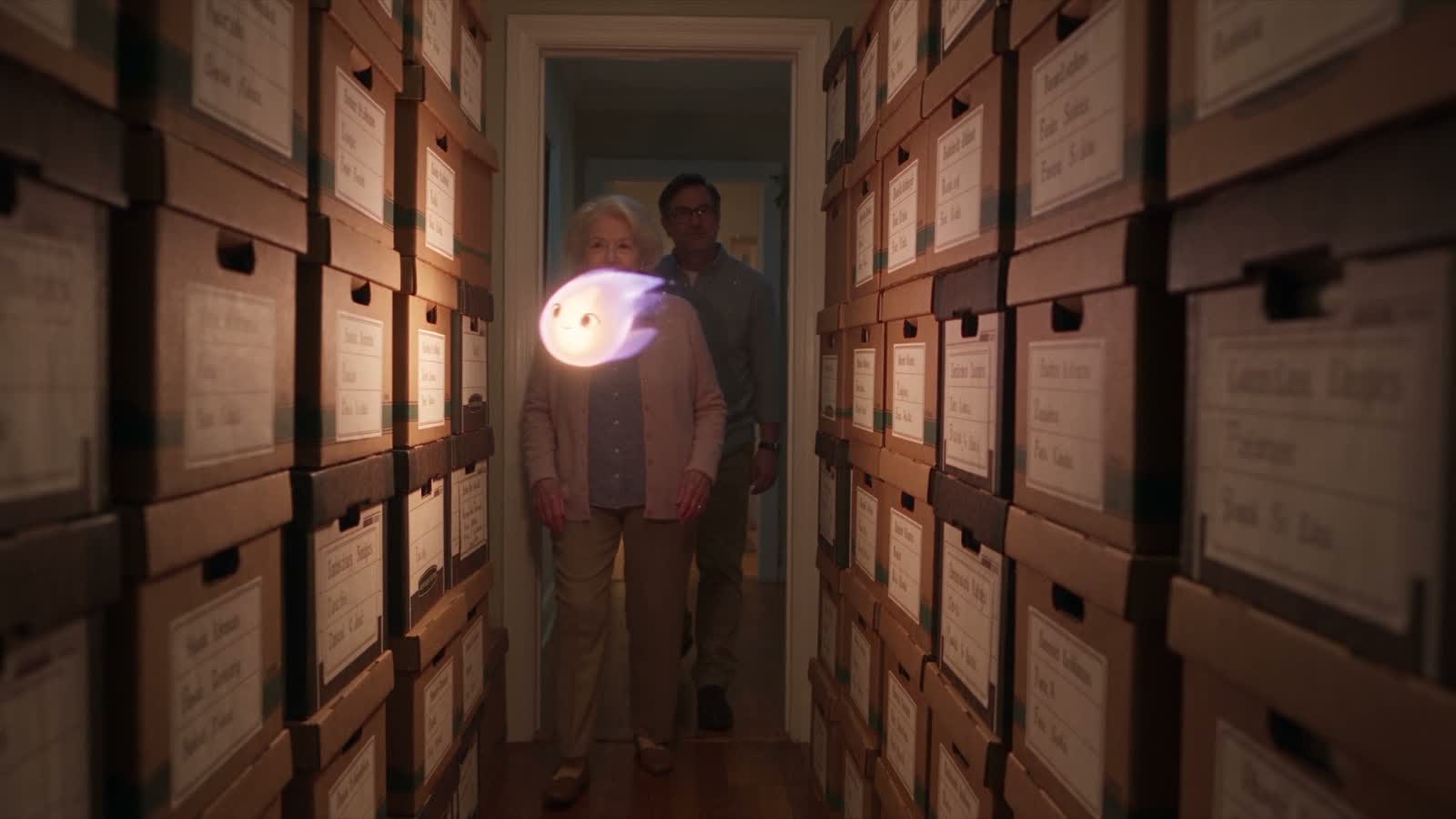 A narrow hallway lined floor to ceiling with labelled archive boxes, a small light leading an older woman and her son through it